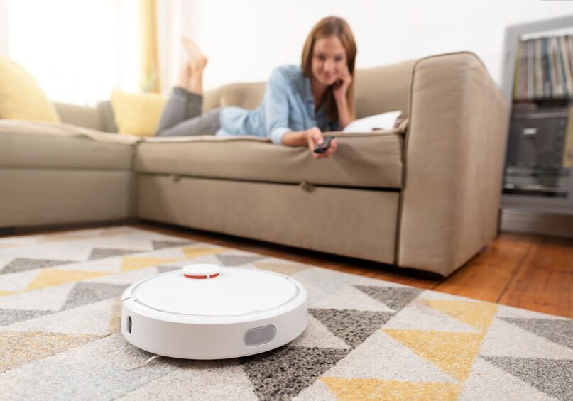 A woman is laying on the couch next to a robot vacuum.